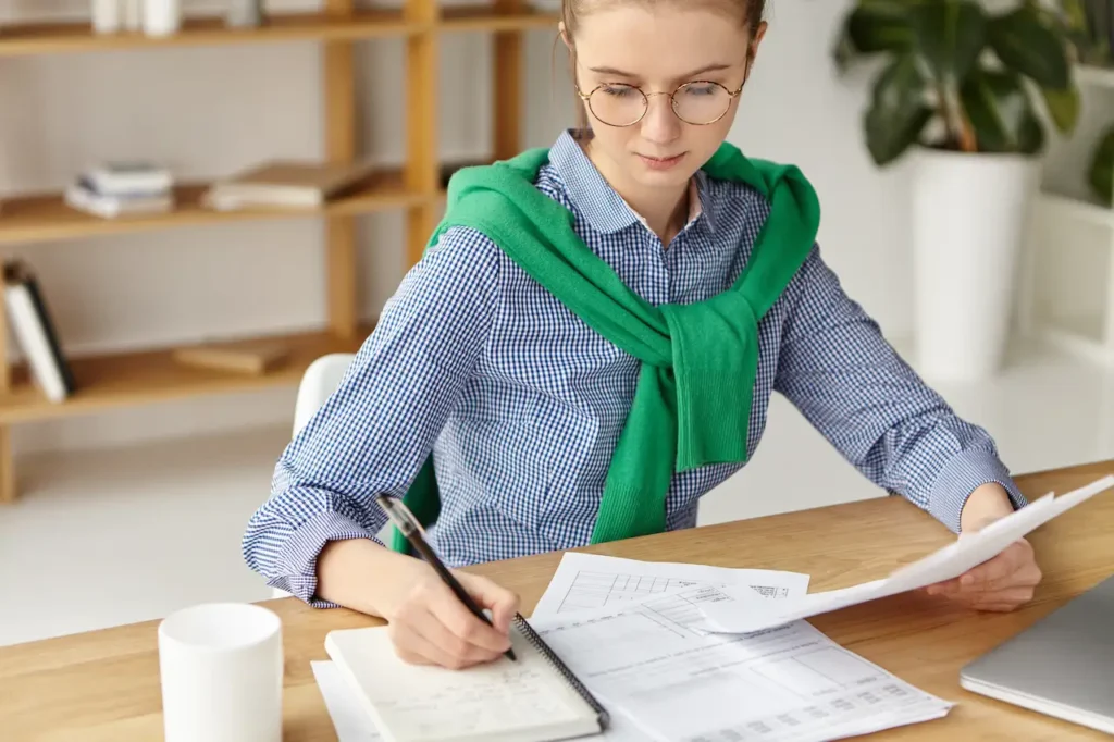 Woman in formal attire with glasses sitting at a desk, writing in a notebook, and reviewing documents. She is wearing a green sweater around her shoulders and has a cup of beverage nearby.