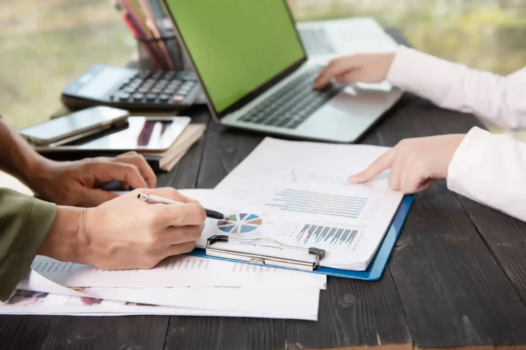Two women sitting on a couch, discussing business charts and graphs. One is holding a tablet while the other holds a clipboard. A laptop, calculator, and office supplies are visible on the table.