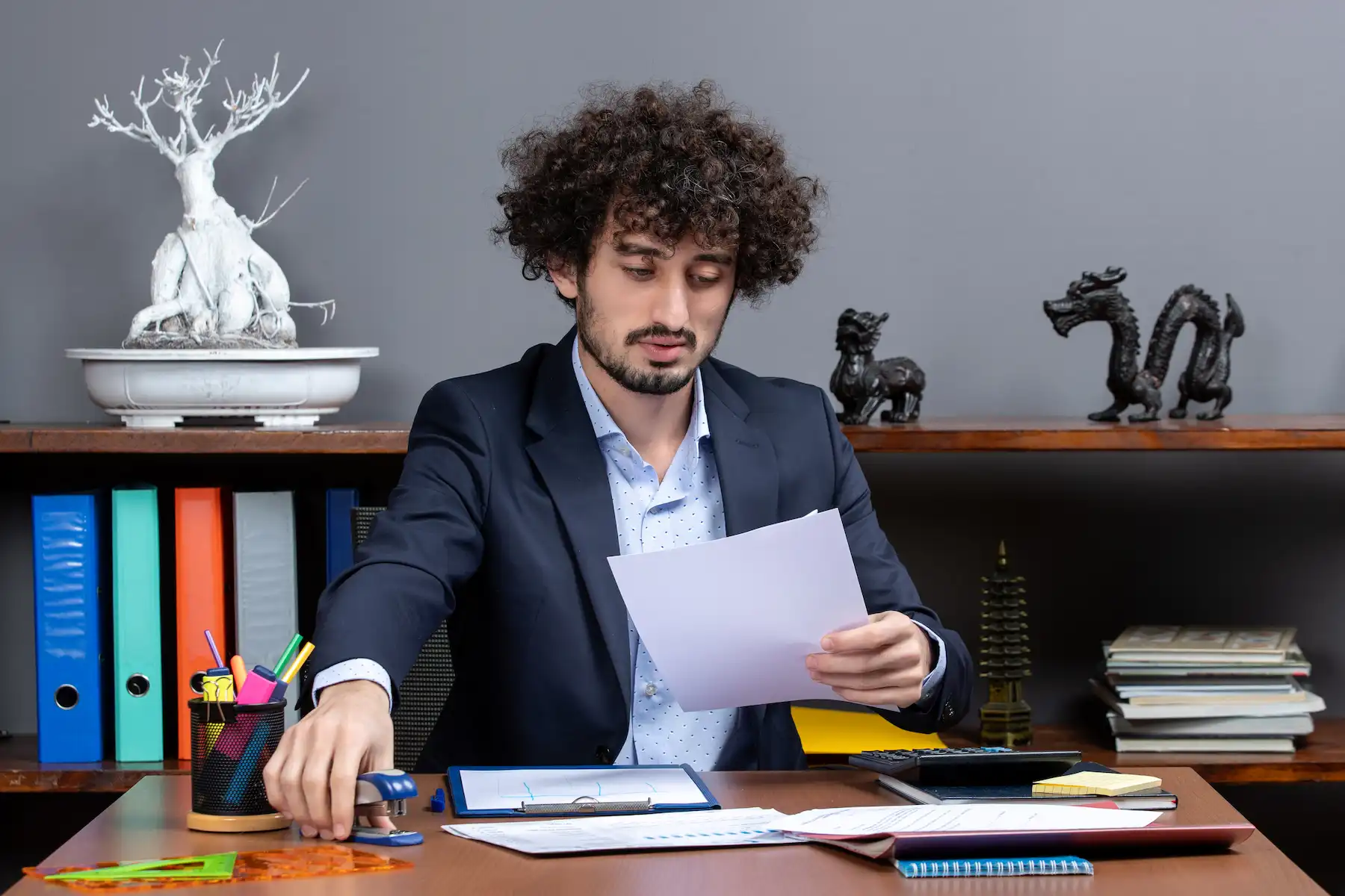 Young businessman working at a desk, holding paper and using a stapler with office supplies in the background.