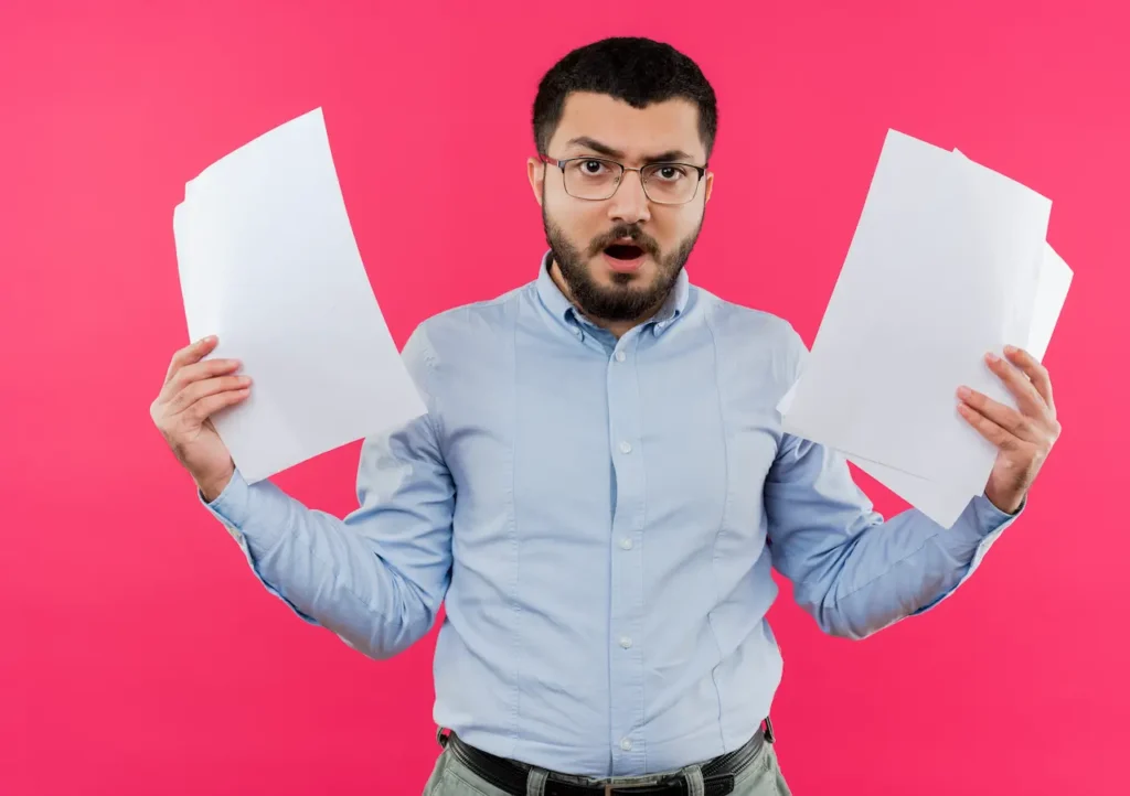 Frustrated young man with a beard and glasses, wearing a blue shirt, holding blank sheets of paper in both hands with an angry expression.