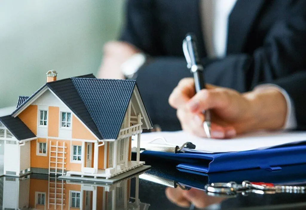 Model house on a table beside a person signing real estate documents with a pen, symbolizing property purchase or agreement.