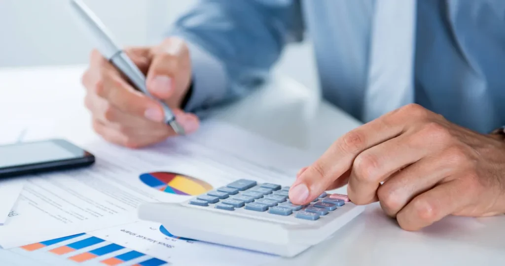 Close-up of a professional using a calculator and reviewing financial charts while taking notes at a desk