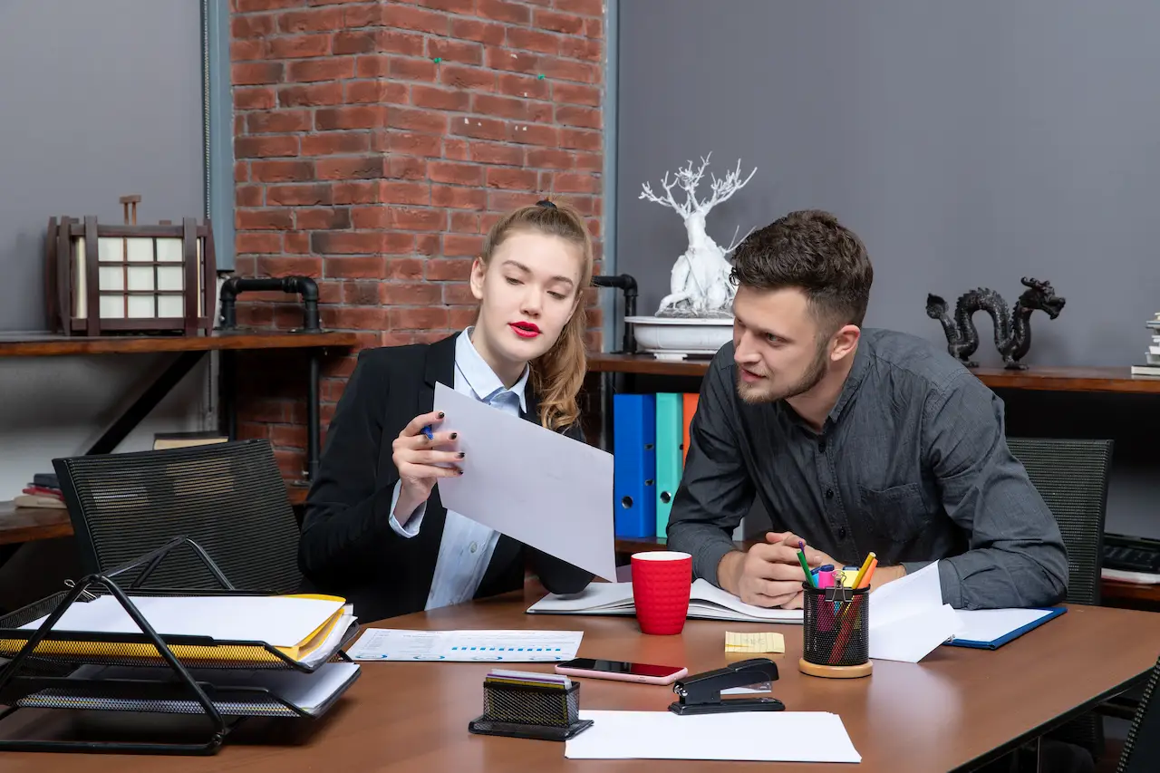Two colleagues in a modern office discussing a document. One is holding the paper and pointing at it while the other is looking at it attentively. There are office supplies like pens, a stapler, and a coffee cup on the desk.