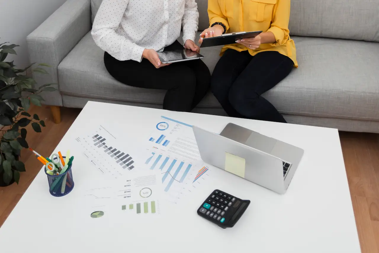 Two women sitting on a couch, discussing business charts and graphs. One is holding a tablet while the other holds a clipboard. A laptop, calculator, and office supplies are visible on the table.