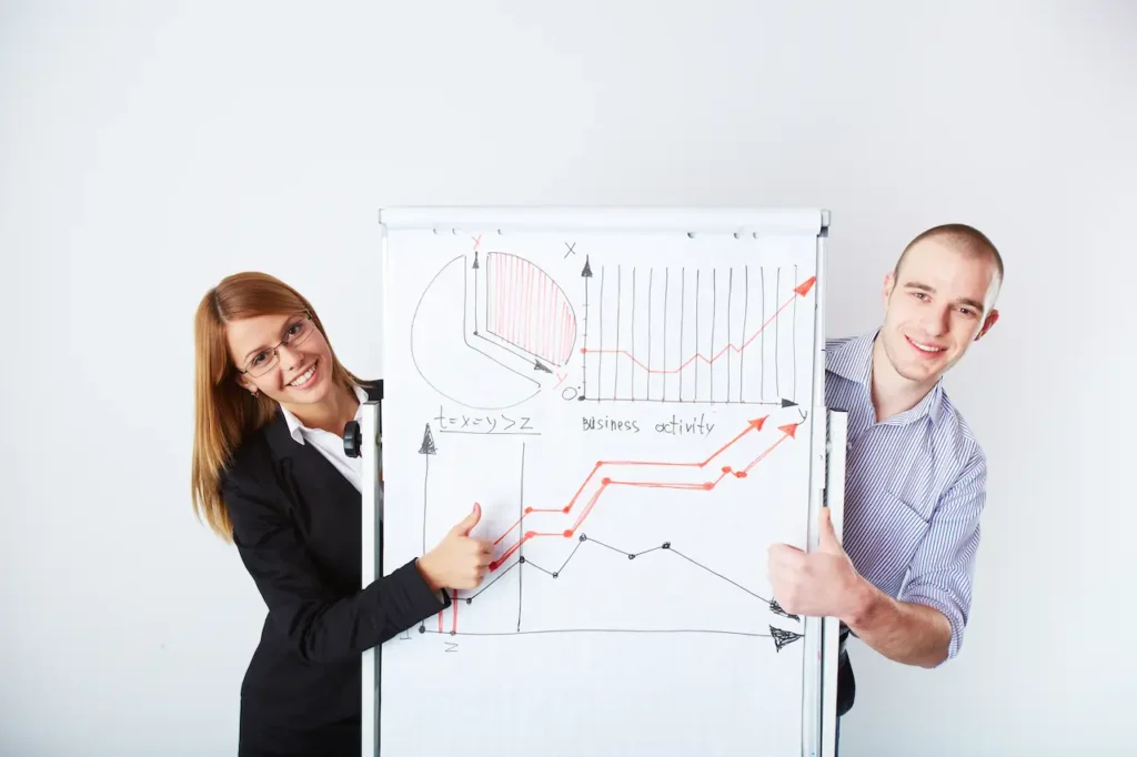 A man and a woman standing next to a whiteboard with graphs and charts, both smiling and giving a thumbs-up to indicate business growth and success.