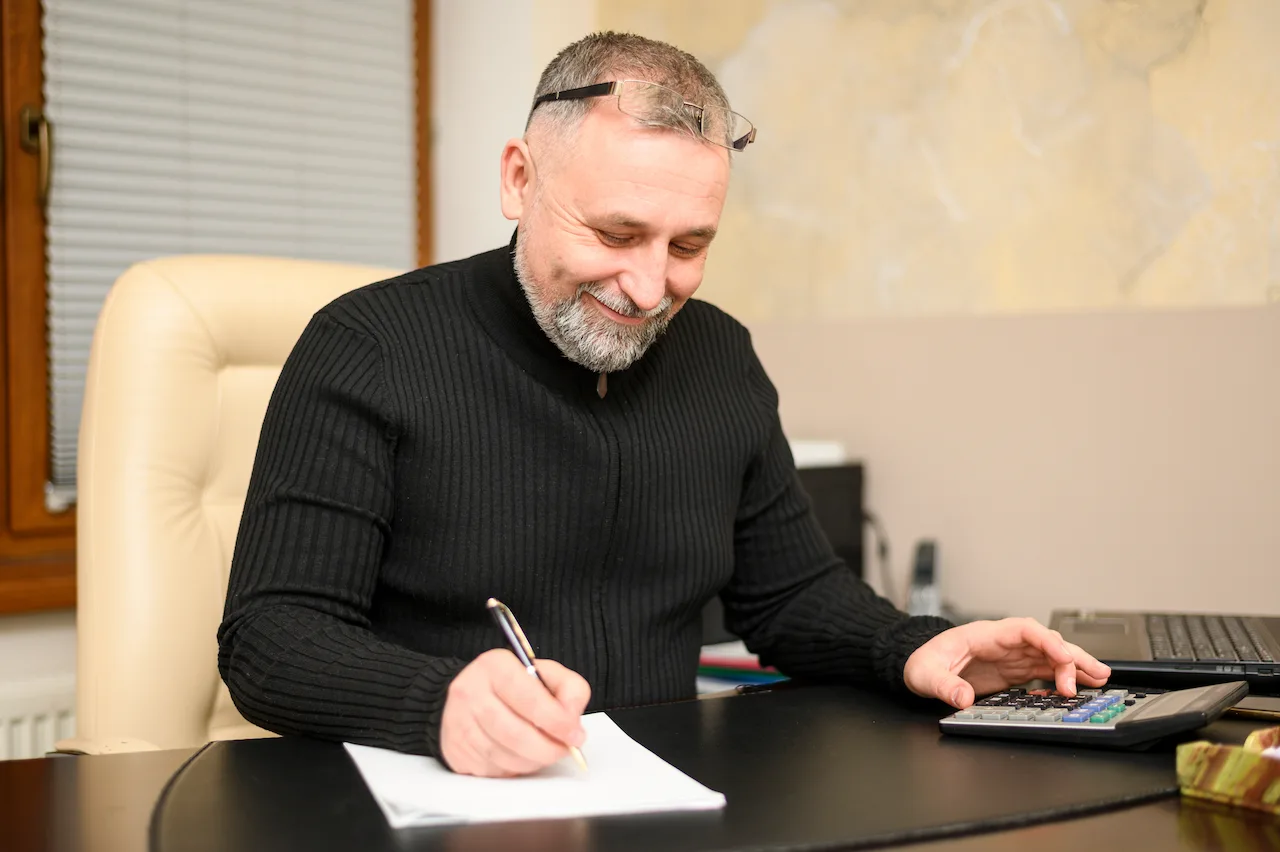 Smiling mature man writing notes at a desk with a pen while using a calculator in a home office